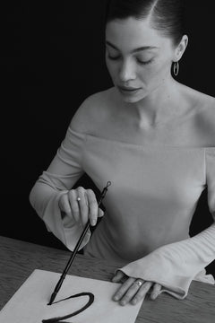 Model practicing Japanese calligraphy in cream dress, Gyosho silver ring visible on the brush hand
