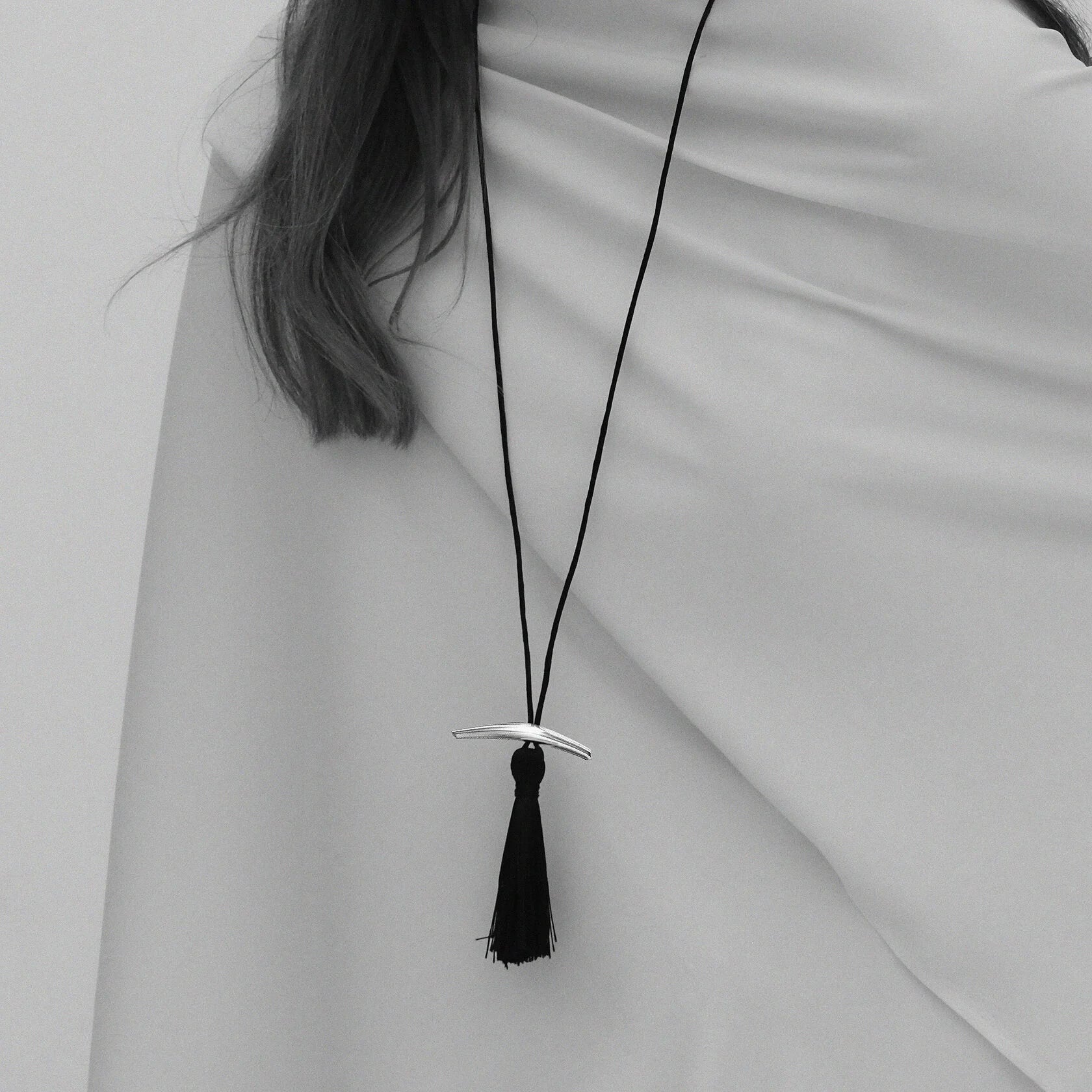 Black-and-white drape close-up of Lera Treyger Dai sterling silver curved bar pendant with black silk tassel on cord
