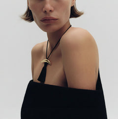 Model in off-shoulder black wool wearing Lera Treyger Dai 14k yellow gold curved bar pendant with long black silk tassel