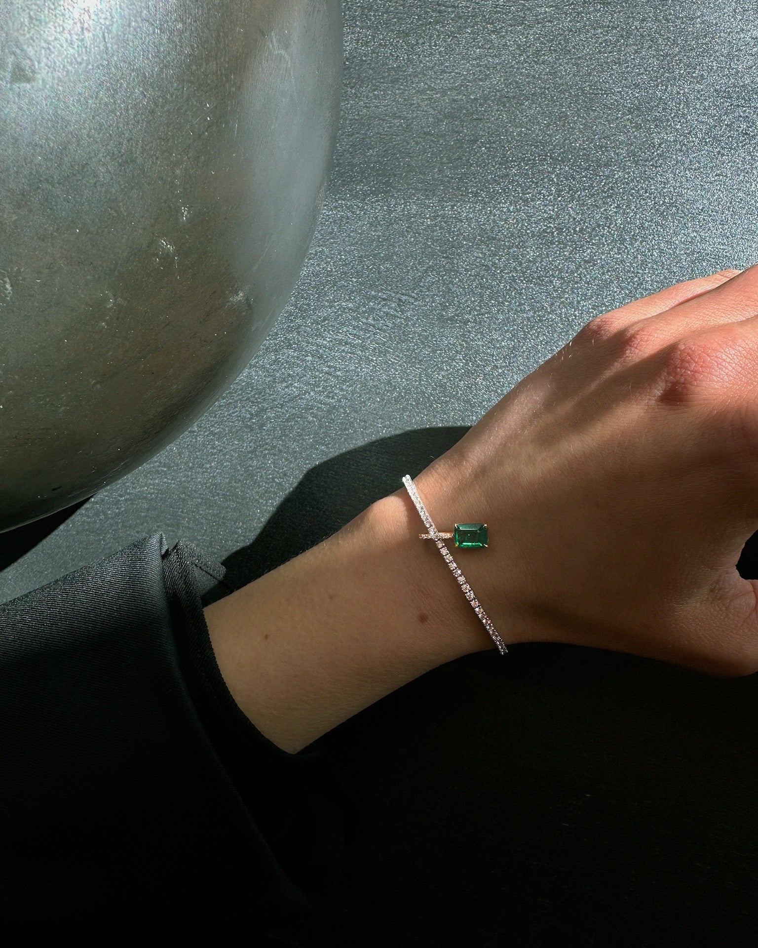 Close-up of Lera Treyger Ukiyo n 3 diamond pave tennis bracelet with emerald-cut emerald accent on hand against grey wall